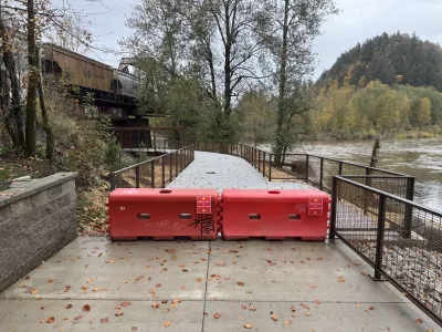 A red barricade blocks access to a trail that curves toward the railroad bridge next to the Sandy River