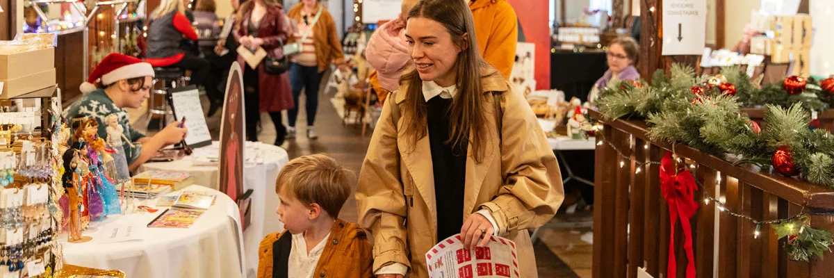 A woman holds a child's hand as they browse gifts at a busy indoor holiday market
